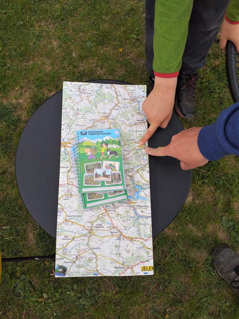na stoliku lezy mapa, dwa palce wskazują na miejscowość Wleń, na mapie leżą dzienniki turystyczne